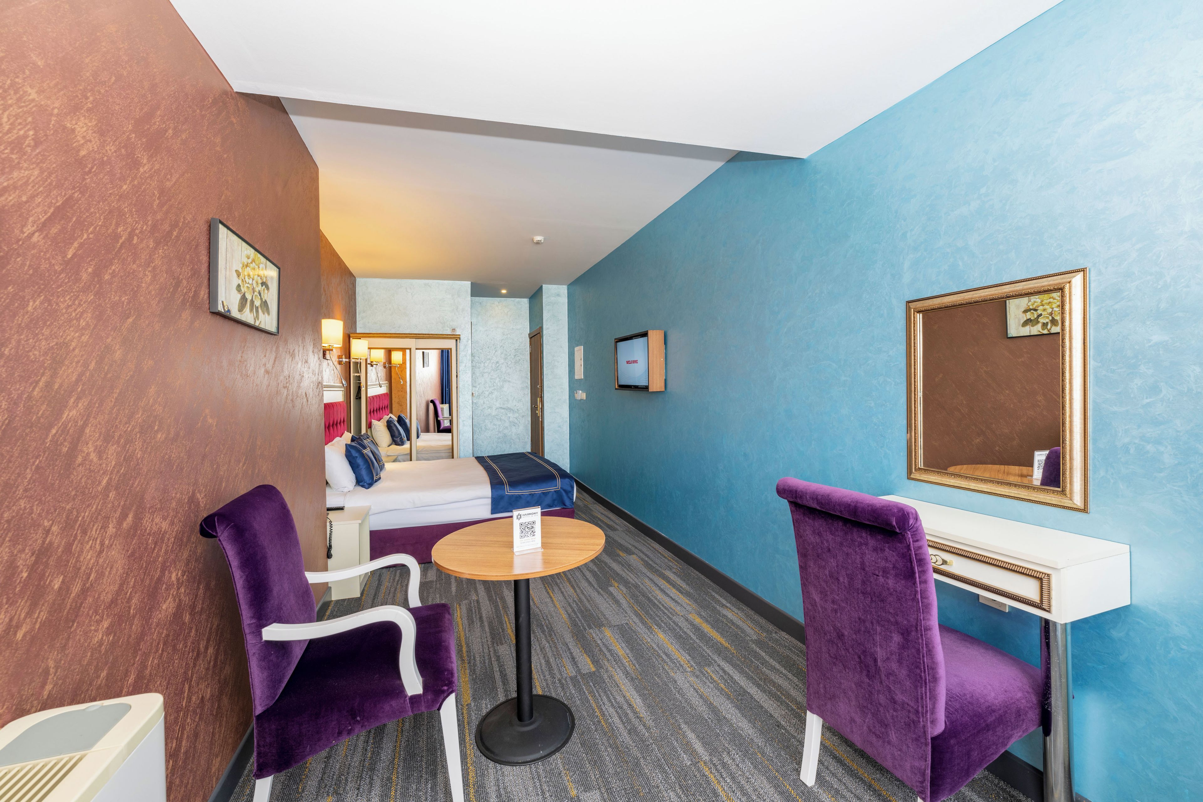 Una habitación de hotel con una cama, dos sillas moradas, una mesita redonda, un escritorio blanco y un espejo.