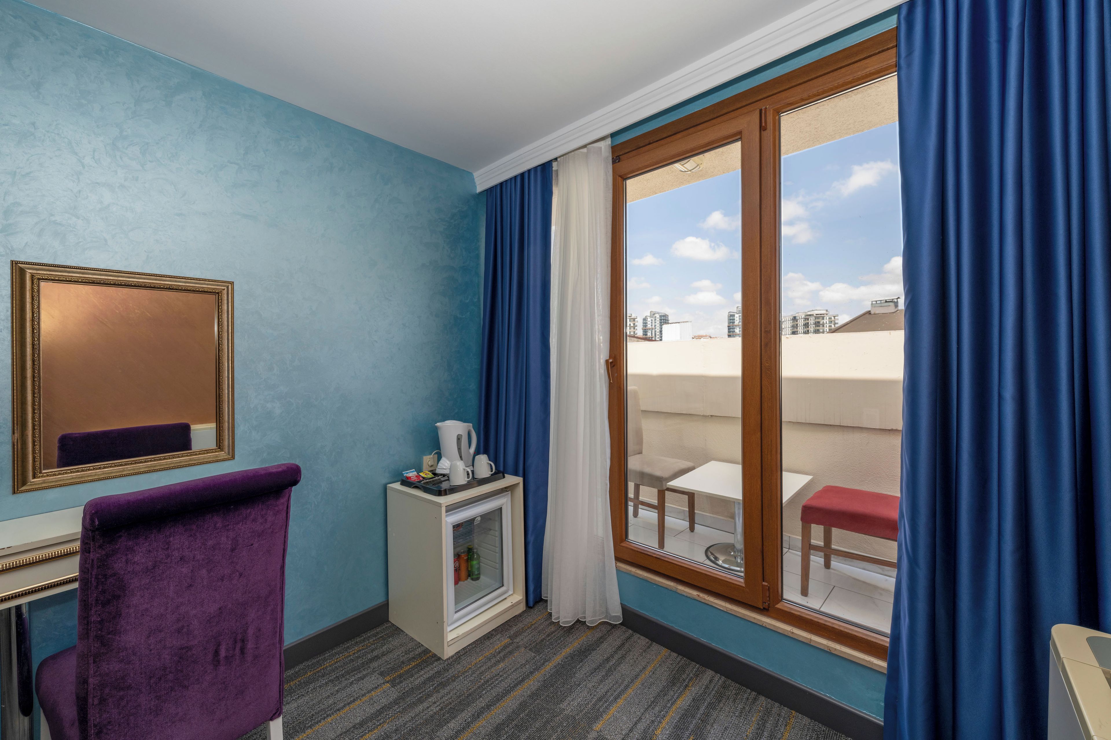 A hotel room with a large sliding glass door to an outdoor balcony, a small minibar, a purple chair, and a mirror on the wall.