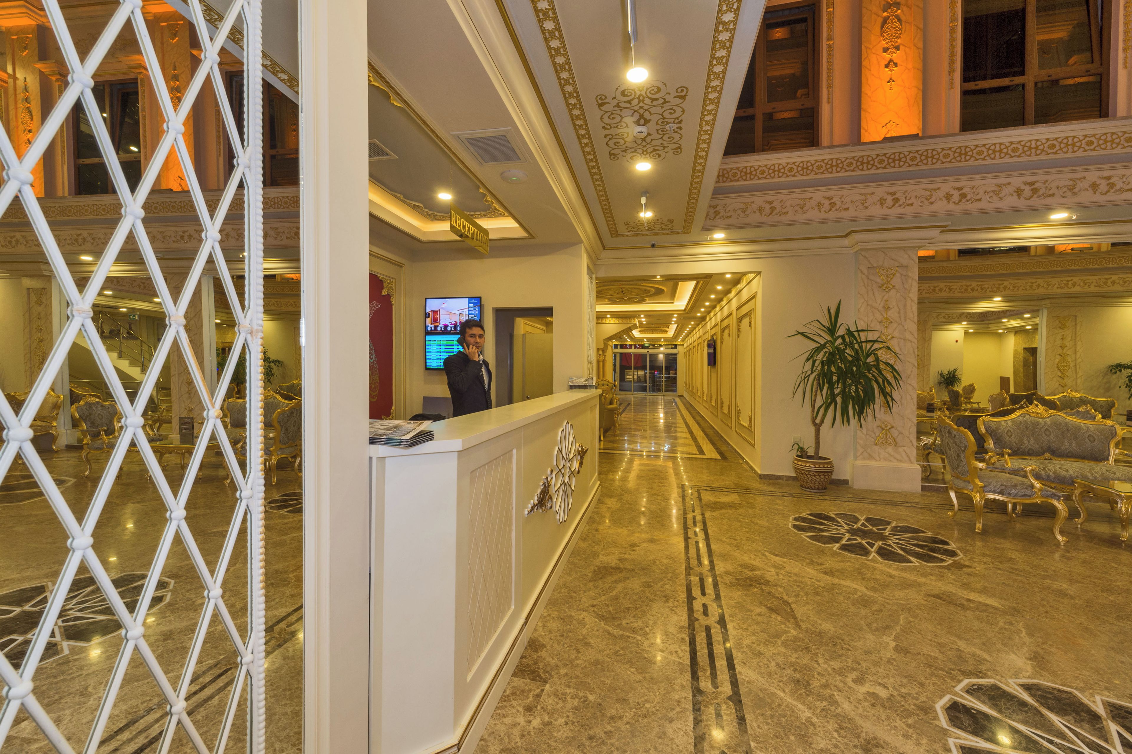 A hotel lobby with a reception desk, a TV screen, a staircase, and ornate wall decorations.