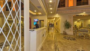 A hotel lobby with a reception desk, a TV screen, a staircase, and ornate wall decorations.