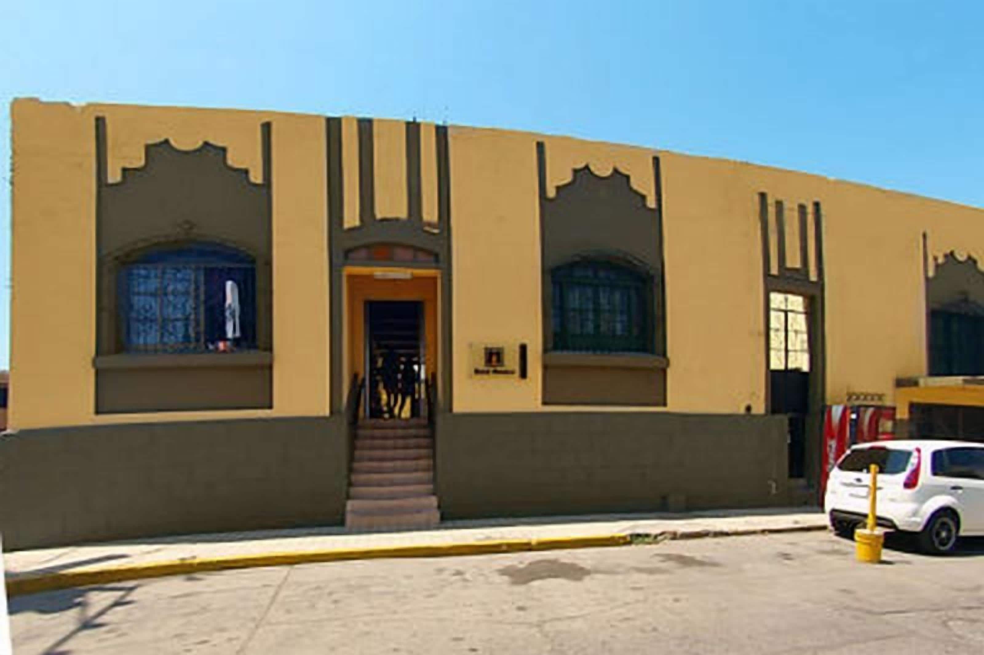 A building with a beige facade, a central entrance with steps, and a small parking area with a white car.