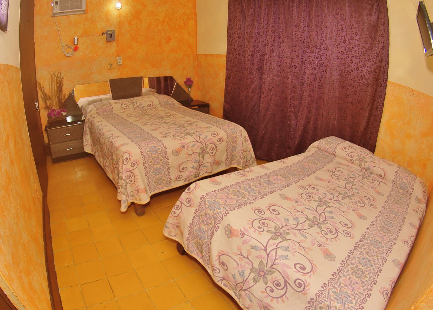 A room with two beds, a nightstand, a vase with flowers, and a curtain.