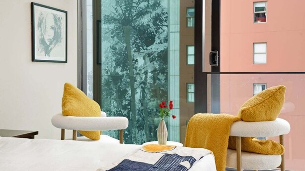 Ein Hotelzimmer mit einem Bett, zwei Stühlen mit gelben Kissen, einer Blumenvase und einem großen Fenster mit Blick auf Bäume und ein Gebäude.