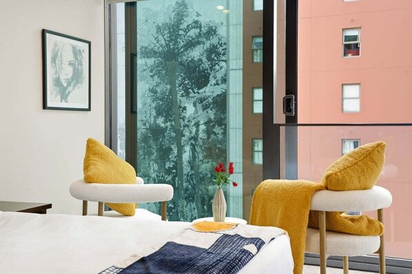 A hotel room with a bed, two chairs with yellow cushions, a vase with flowers, and a large window with a view of trees and a building. - Stylish Apartment 130m to Int Conference Centre (Auckland CBD)