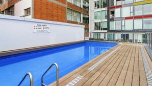 Ein Poolbereich mit einem Sicherheitsschild, einer Holzterrasse und einem modernen Gebäude mit vielen Fenstern.