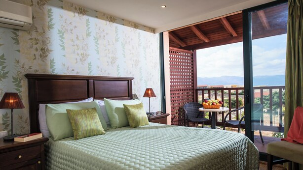A hotel room with a bed, a nightstand, a lamp, a window with a view, and a balcony with a table and chairs.