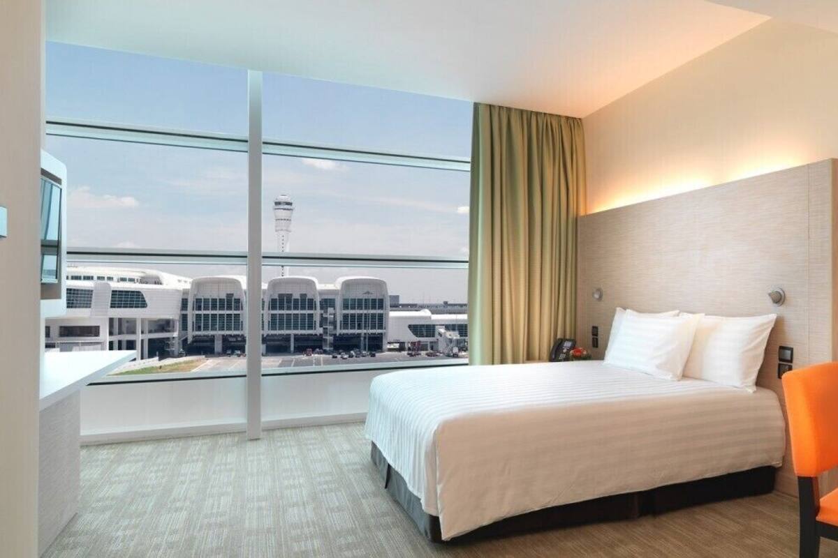 A hotel room with a large bed, a desk, and a chair. There is a window with a view of airport buildings and a tower.