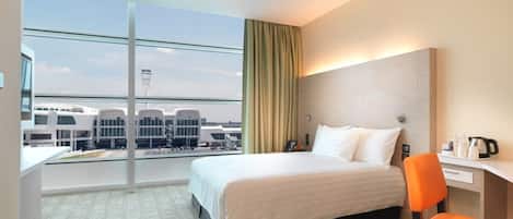 Une chambre d’hôtel avec un grand lit, un bureau et une chaise. Il y a une fenêtre donnant sur les bâtiments de l’aéroport et une tour.