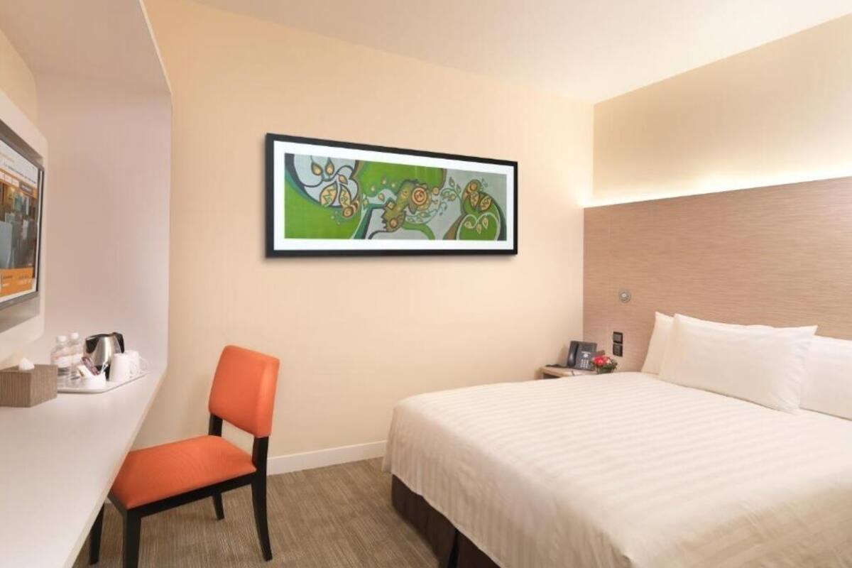 A hotel room with a bed, a desk with a chair, a painting on the wall, and a nightstand with a lamp.