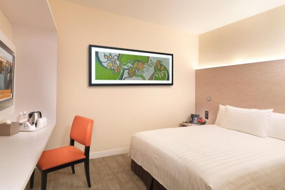 A hotel room with a bed, a desk with a chair, a painting on the wall, and a nightstand with a lamp.