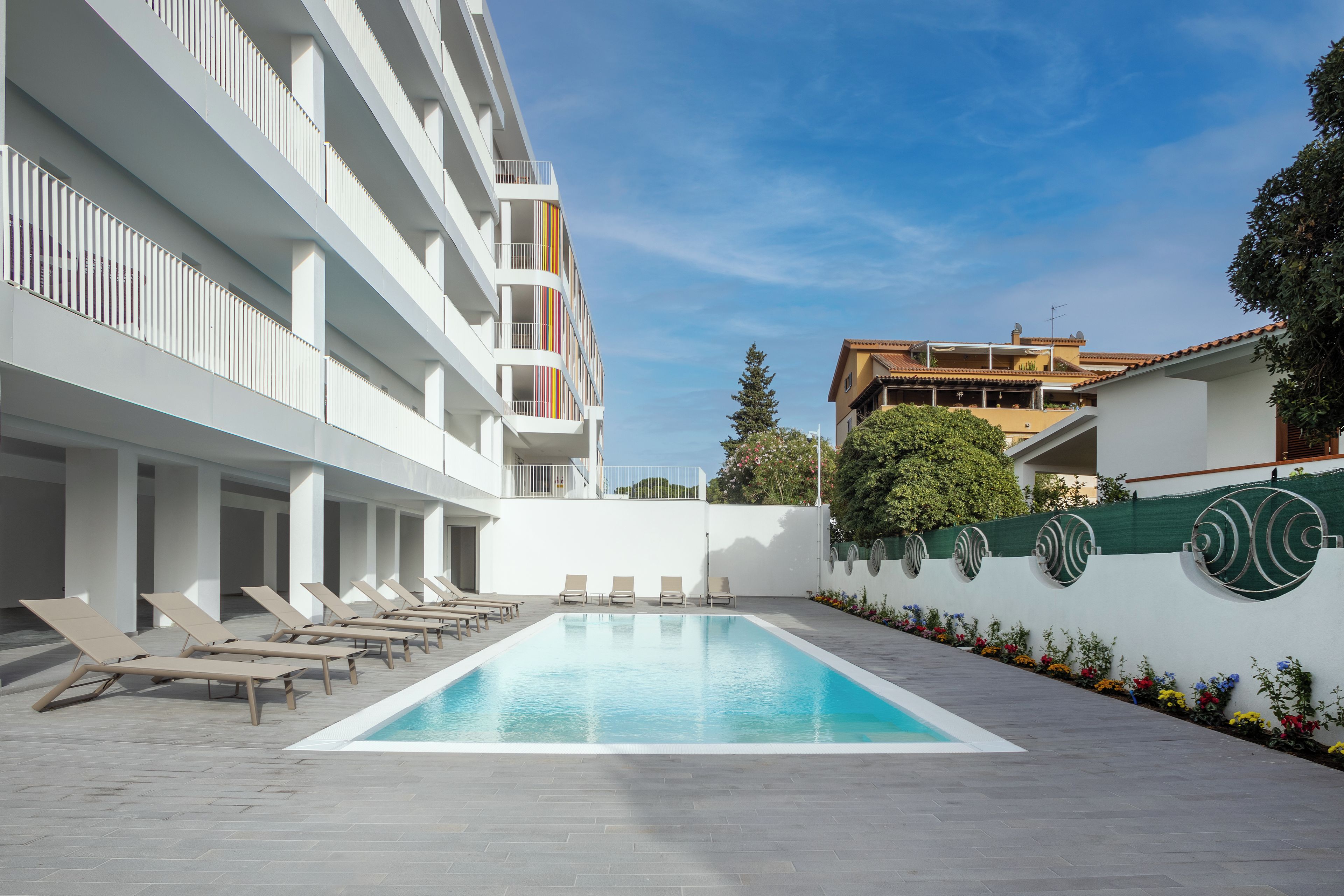 A swimming pool with lounge chairs, a white building with balconies, and a building with a tiled roof.