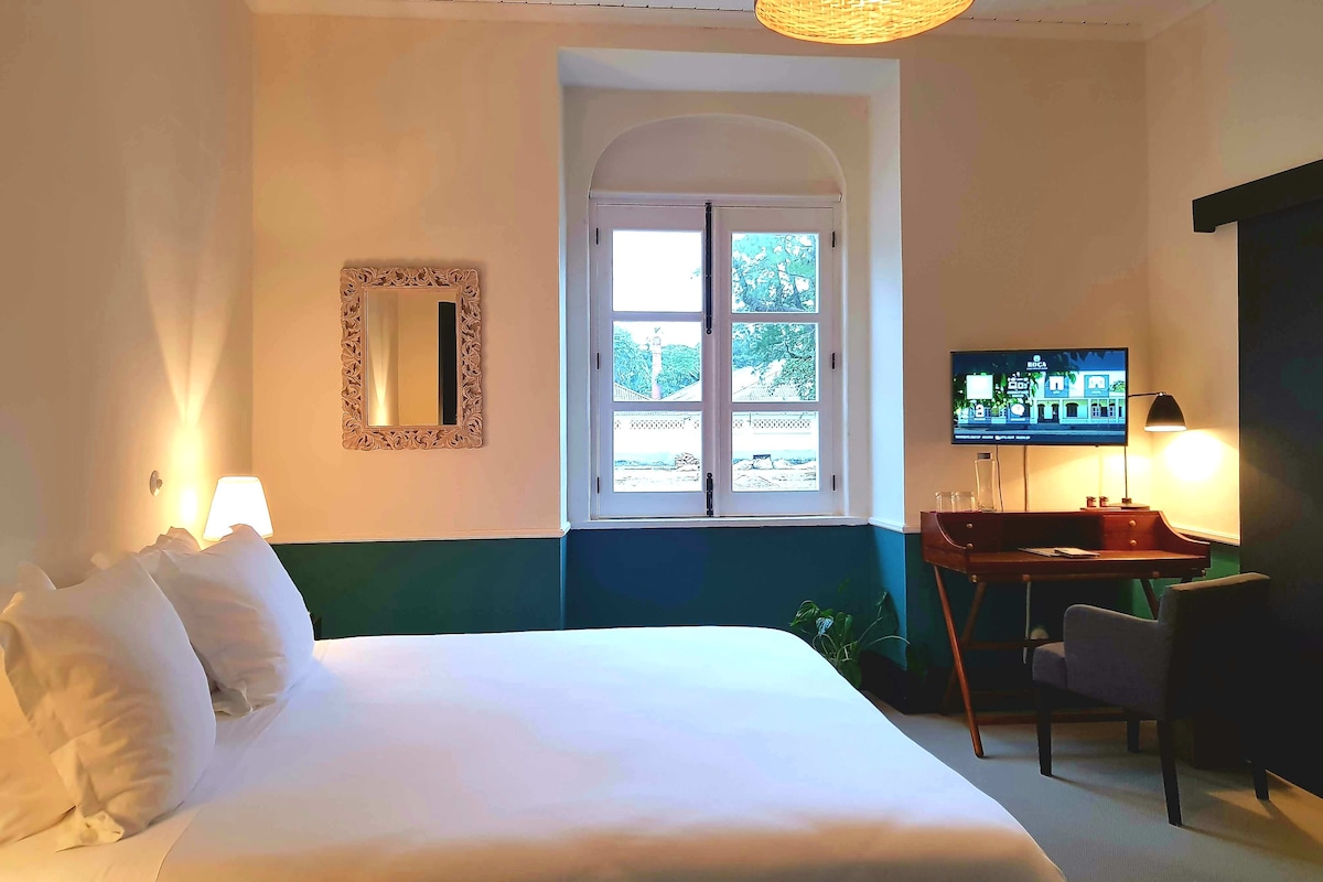A hotel room with a bed, a television, a desk with a chair, a window with a view, and a mirror on the wall.