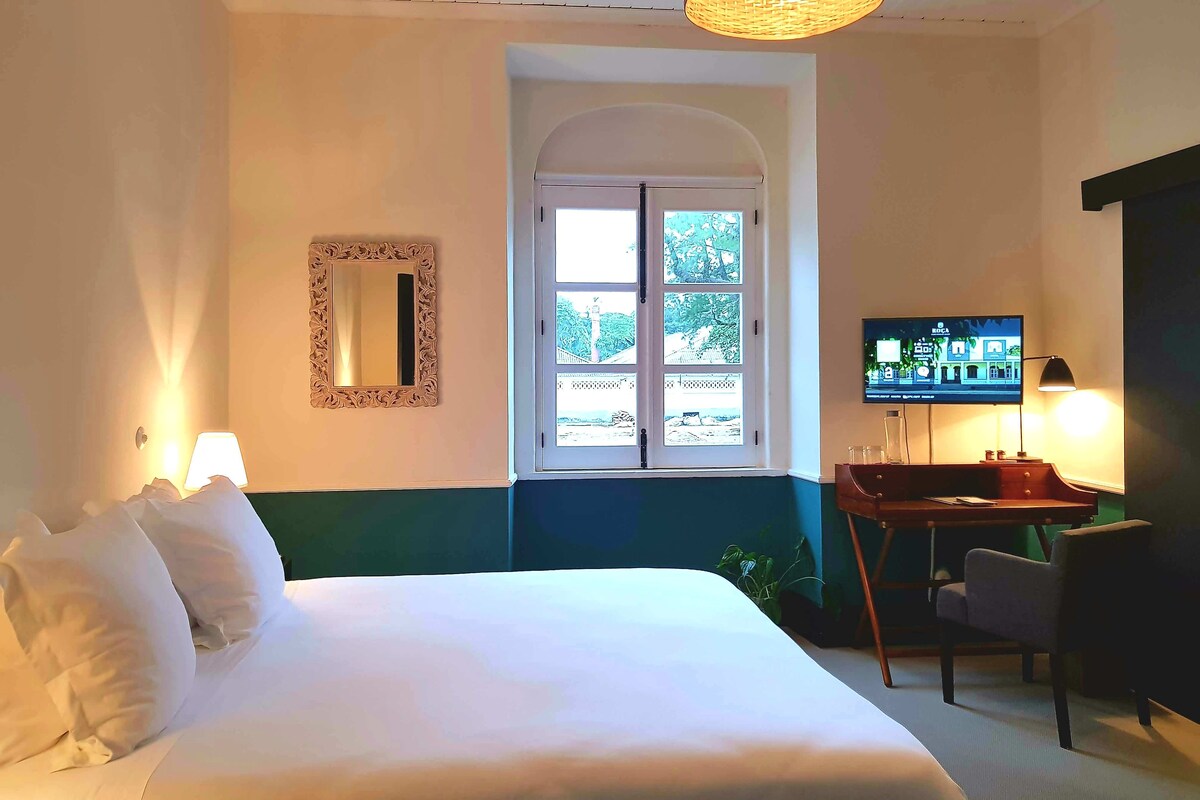 Habitación de hotel con cama, televisión, escritorio con silla, ventana con vistas y espejo en la pared.