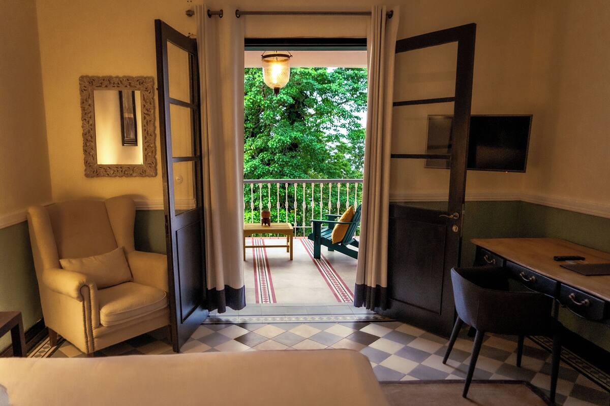 A hotel room with a bed, a desk, a chair, a sofa, and a balcony with a view of trees.