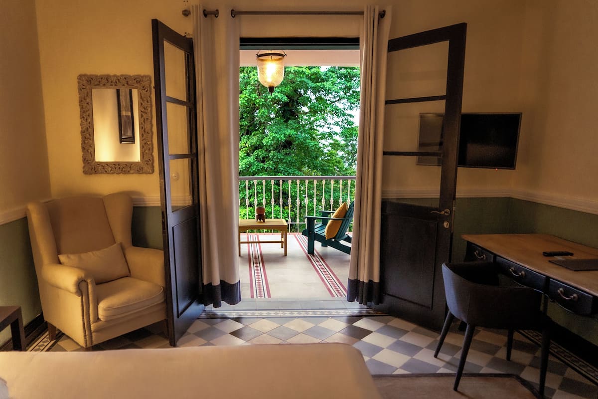 A hotel room with a bed, a desk, a chair, a sofa, and a balcony with a view of trees.