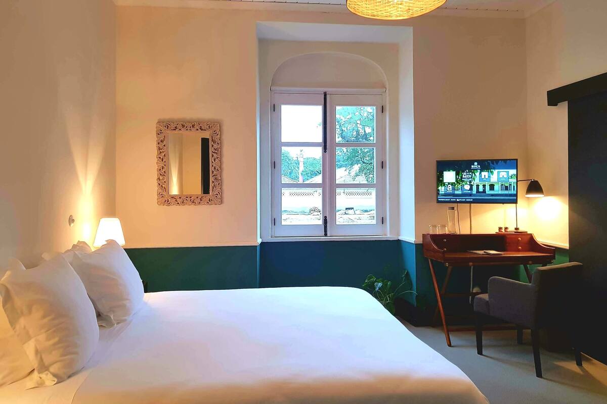 A hotel room with a bed, a television, a desk with a chair, a window with a view, and a mirror on the wall.