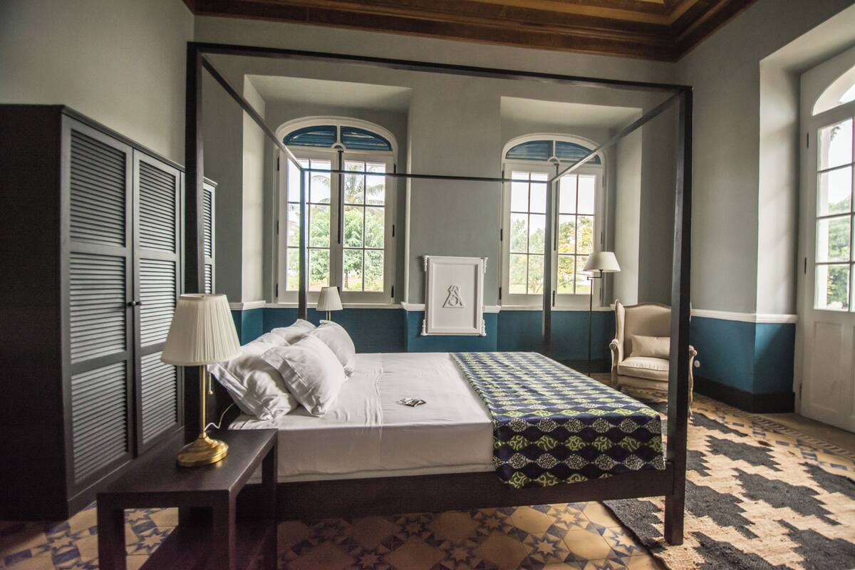 A four-poster bed with a white comforter and pillows, a bedside lamp, and a patterned throw.