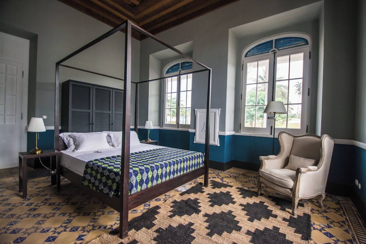 A four-poster bed with a patterned bedspread, a chair, and a large window with shutters.