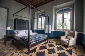 A four-poster bed with a patterned bedspread, a chair, and a large window with shutters.