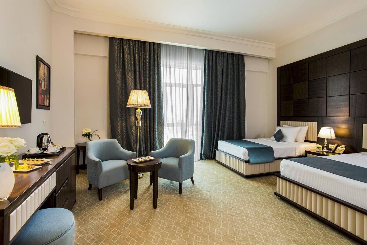 Quarto de hotel com uma cama, duas poltronas, uma mesa pequena, escrivaninha com luminária e uma janela grande com cortinas.