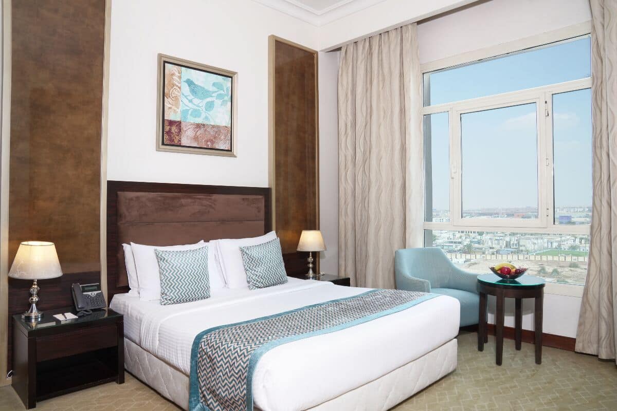 Quarto de hotel com uma cama grande, uma mesa com luminária, uma cadeira, uma janela com cortinas e vista para a cidade.