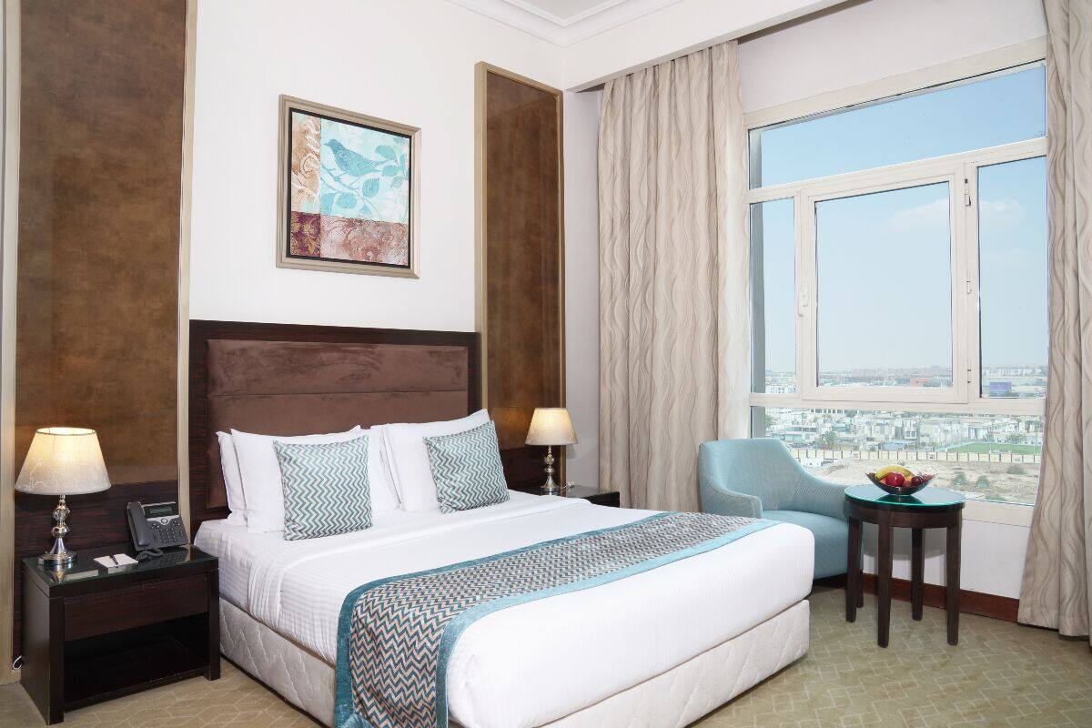 Quarto de hotel com uma cama grande, uma mesa com luminária, uma cadeira, uma janela com cortinas e vista para a cidade.