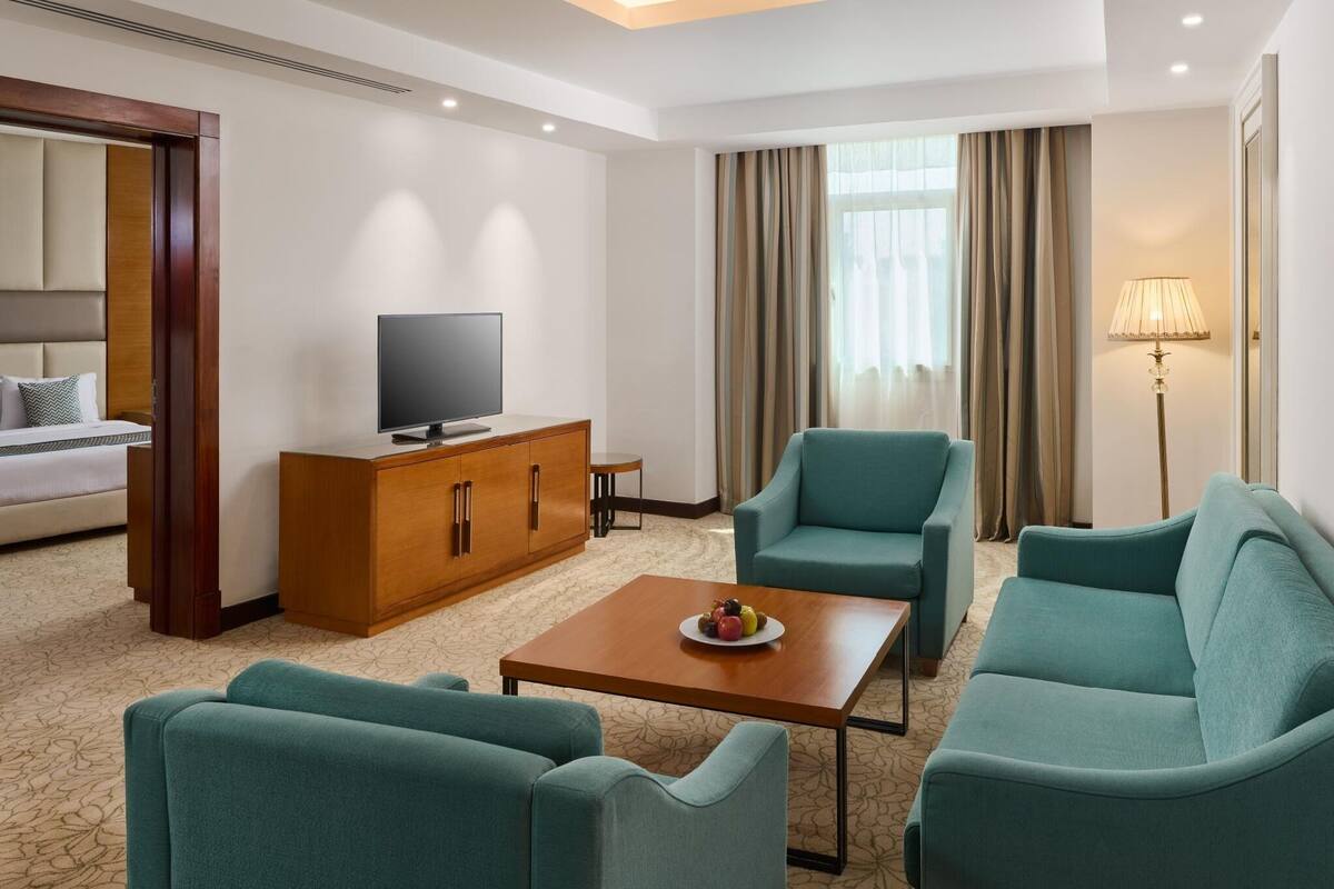 Quarto de hotel com uma cama, televisão, aparador de madeira, mesa de centro e poltronas azul-turquesa.