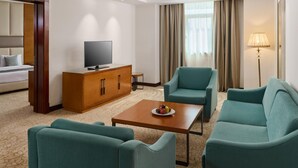 A hotel room with a bed, a television, a wooden cabinet, a coffee table, and teal armchairs. - Gewan Hotel Cairo (Giza)