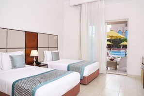 A hotel room with two beds, a nightstand with a lamp, a view of a pool and umbrellas outside. - Gewan Hotel Cairo (Giza)
