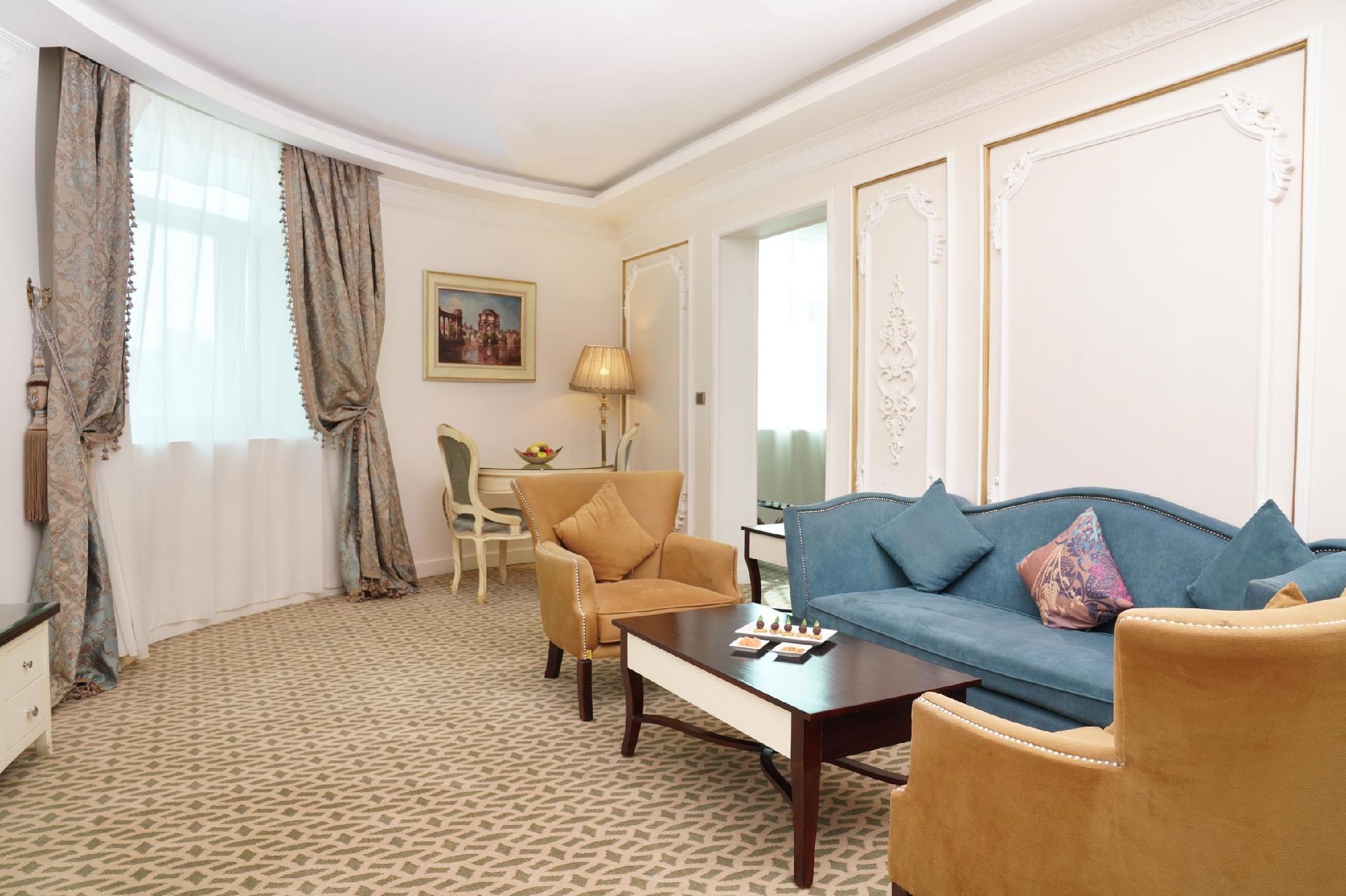Quarto de hotel com um sofá azul, uma poltrona bege, uma mesa de jantar e um tapete estampado.