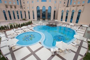 Ein großer Außenpool mit Liegestühlen und Sonnenschirmen, umgeben von einem Hotelgebäude mit Bogenfenstern.