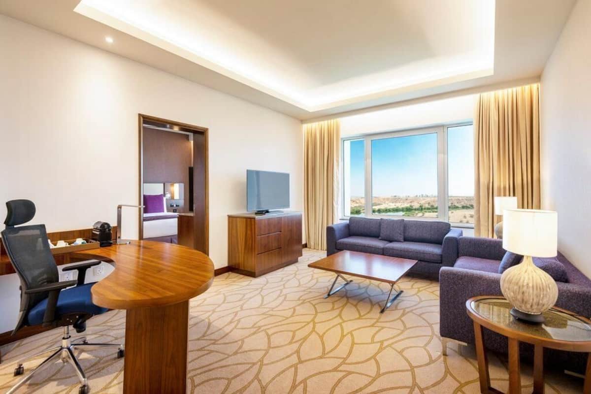 Habitación de hotel con escritorio de conferencias, sofá, televisor y ventana grande con vistas al desierto.