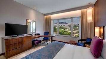 A hotel room with a large bed, a desk with a computer, a TV, a view of a building through the window, and a cityscape.