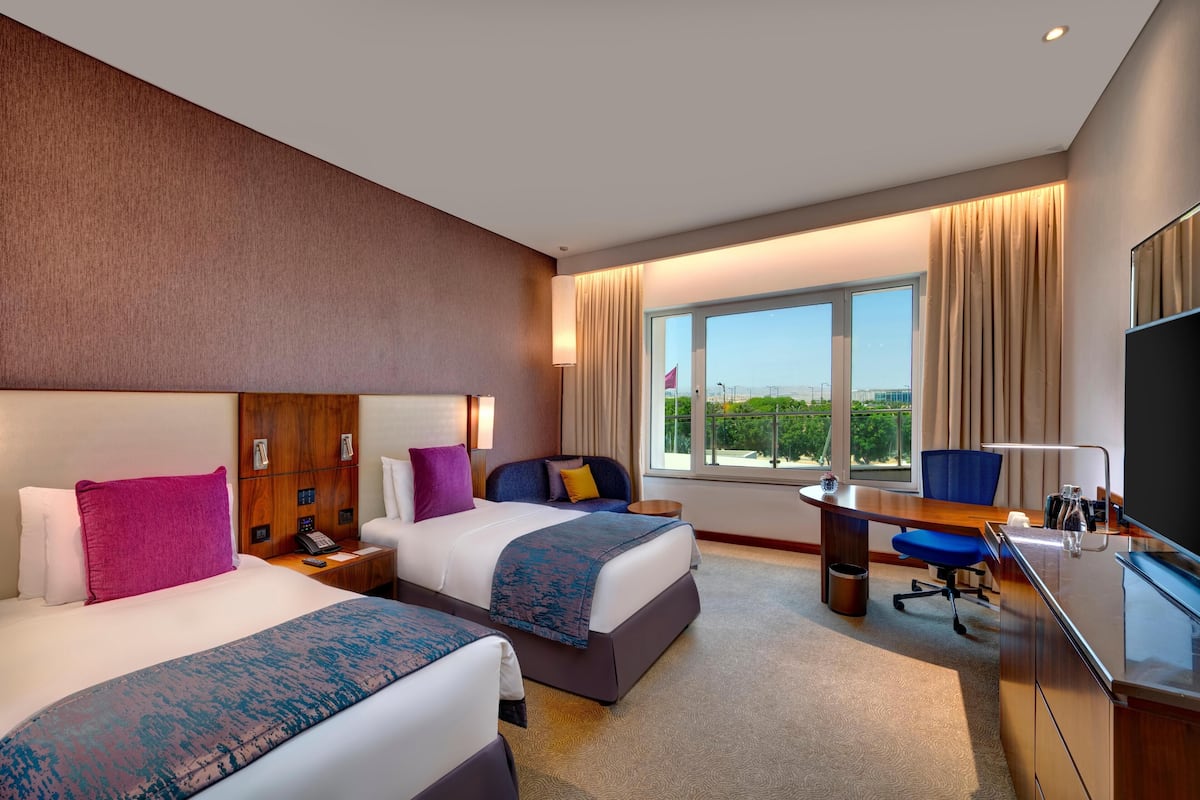 Habitación de hotel con dos camas, un escritorio, una silla, un televisor y un ventanal con vistas.