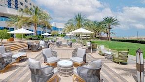 An outdoor seating area with wicker furniture and umbrellas, palm trees, and a building with large windows in the background.
