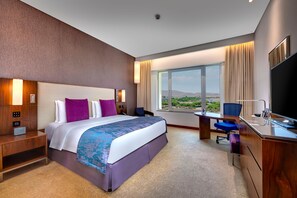A hotel room with a large bed, a desk with a chair, a TV, and a window with a view. - Crowne Plaza Muscat Ocec (Muscat)