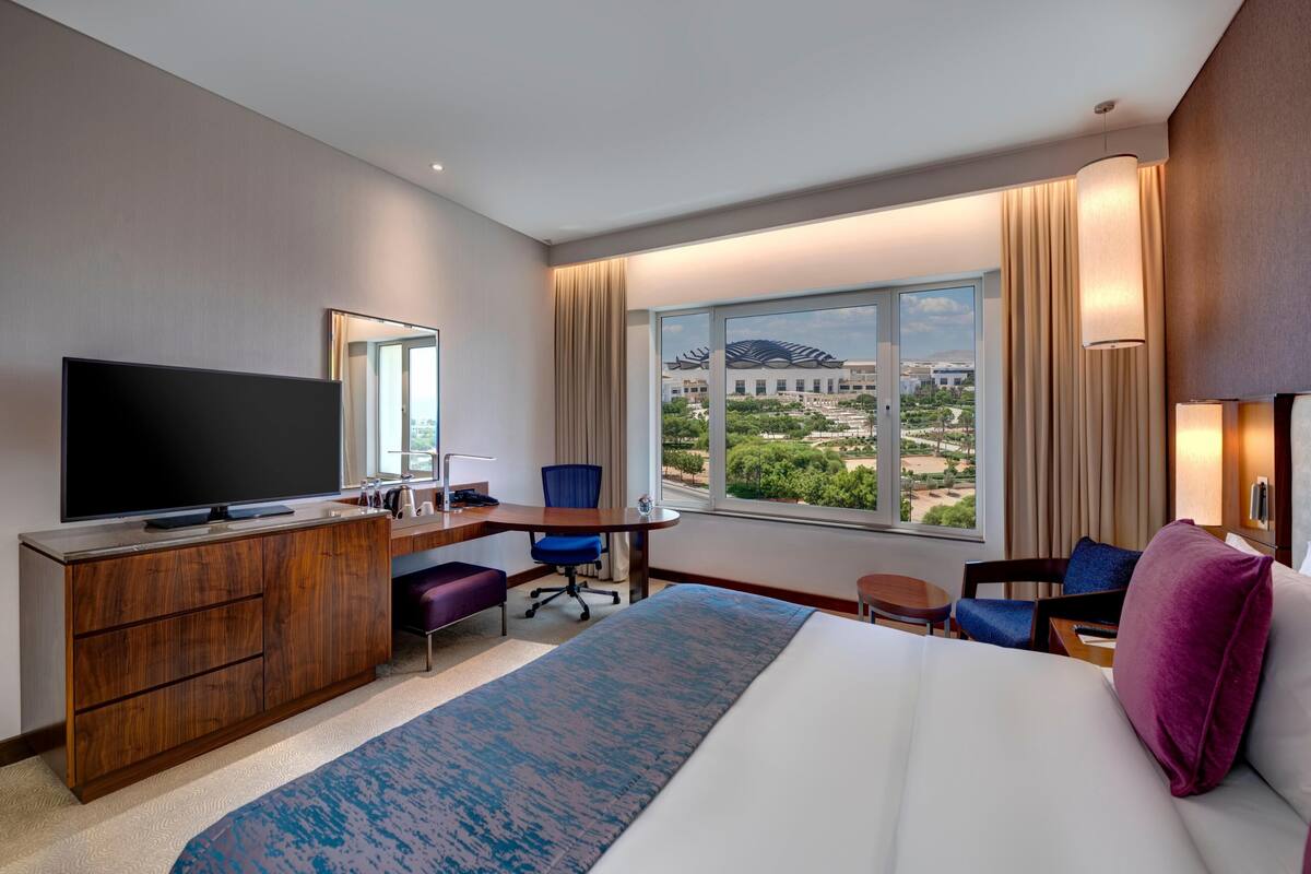Habitación de hotel con una cama grande, un escritorio con ordenador, televisor, vista a un edificio a través de la ventana y un paisaje urbano.