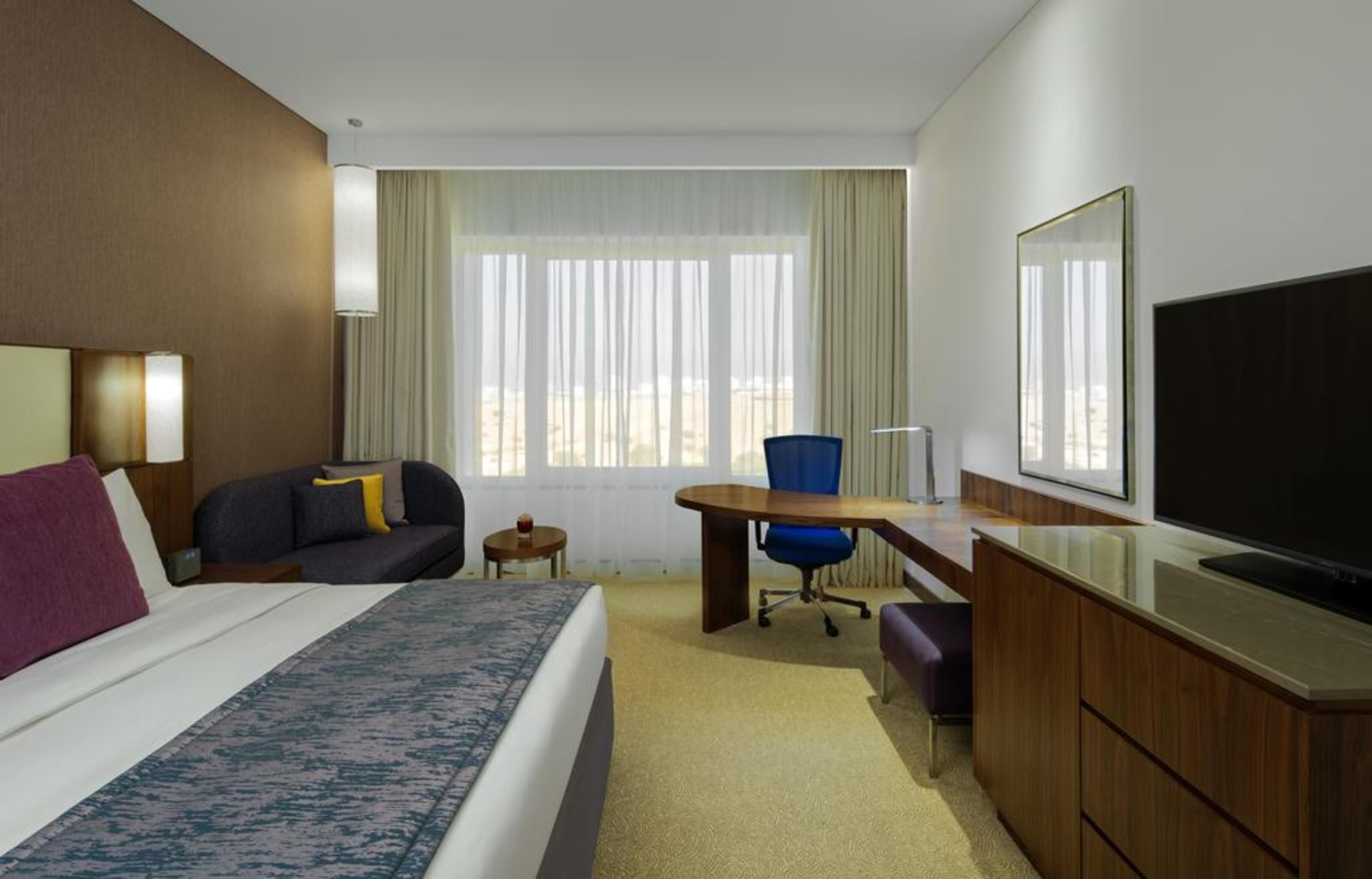 Habitación de hotel con cama, escritorio, silla, televisor y ventana con cortinas.