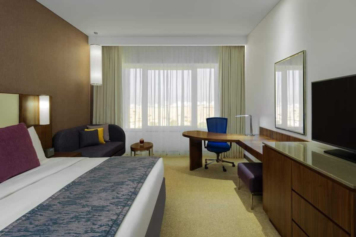 Habitación de hotel con cama, escritorio, silla, televisor y ventana con cortinas.