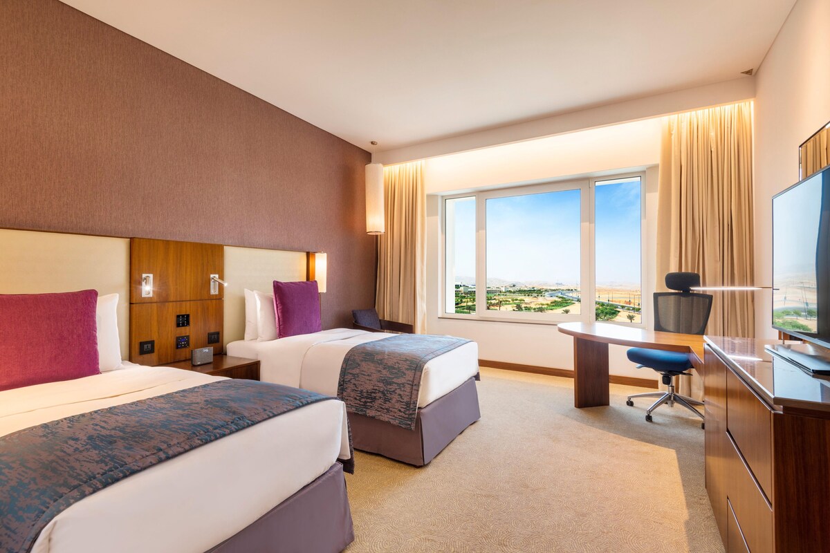 A hotel room with two beds, a desk, a chair, a TV, and a large window with a view.
