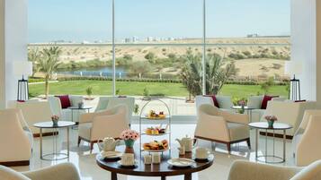 A room with a large window overlooking a beach and cityscape, furnished with chairs, a table, and a tiered serving tray with food.