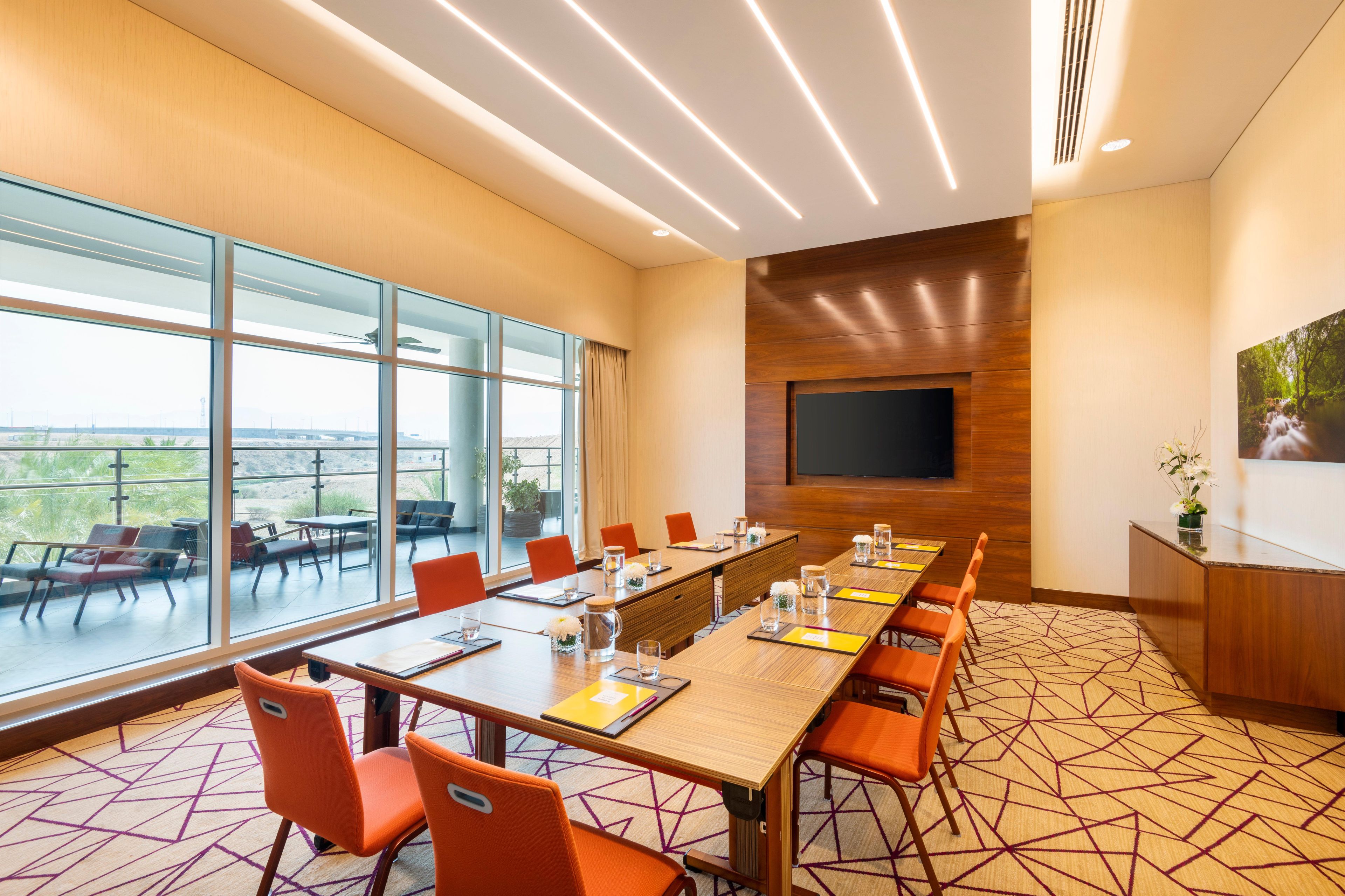 Un salón de conferencias con una larga mesa, sillas anaranjadas, un televisor de pantalla plana y amplias ventanas con vistas.