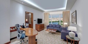 A hotel room with a dining table, sofa, TV, and a view of a body of water. - Crowne Plaza Muscat Ocec (Muscat)