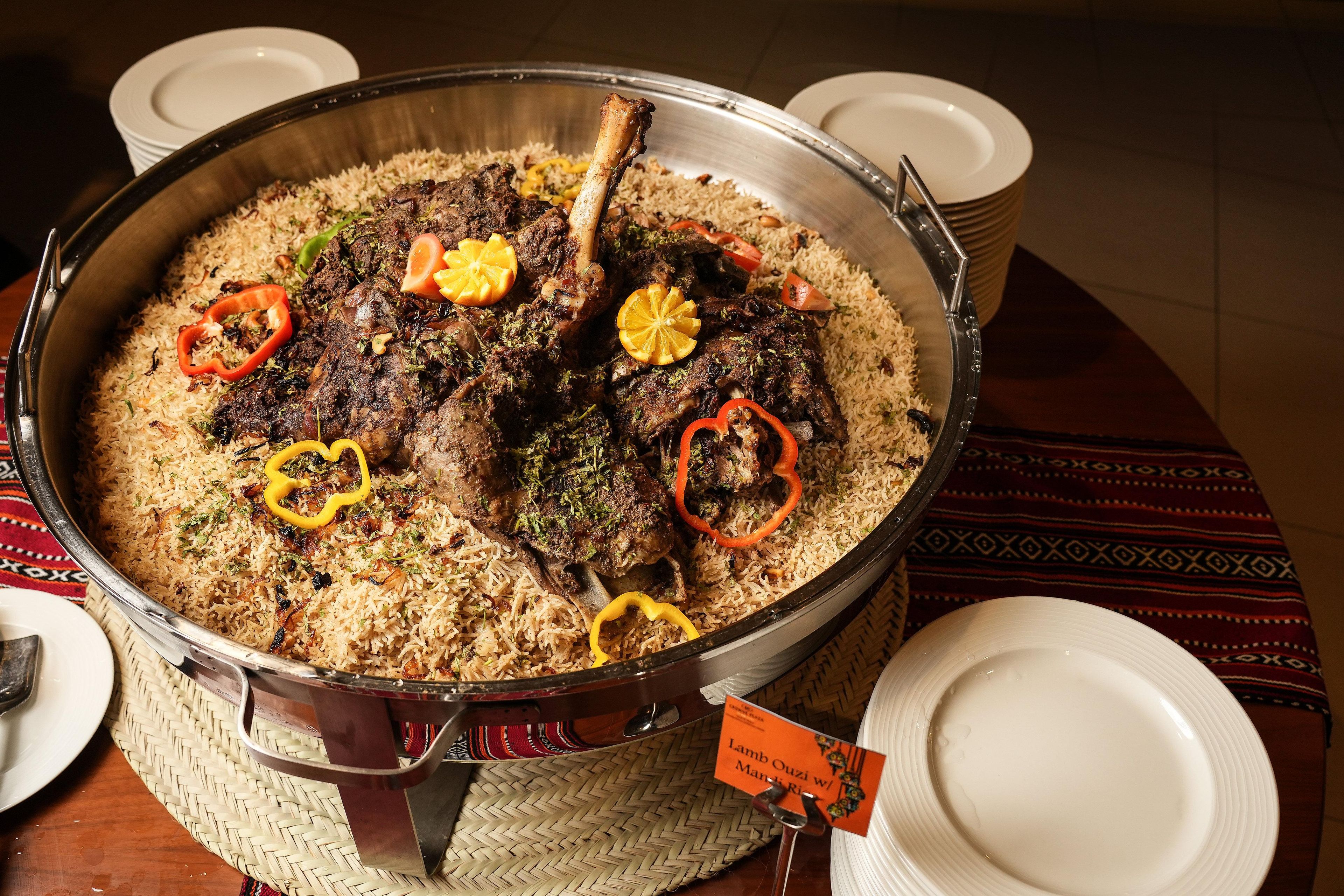 Een groot metalen serveerschaal gevuld met een rijstgerecht, versierd met plakjes rode en gele paprika, en een tag met de tekst 'Lamb Ouzie w/ Marjolee'.