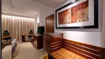 A hotel room with a bed, desk, chair, television, wardrobe, and artwork on the wall.