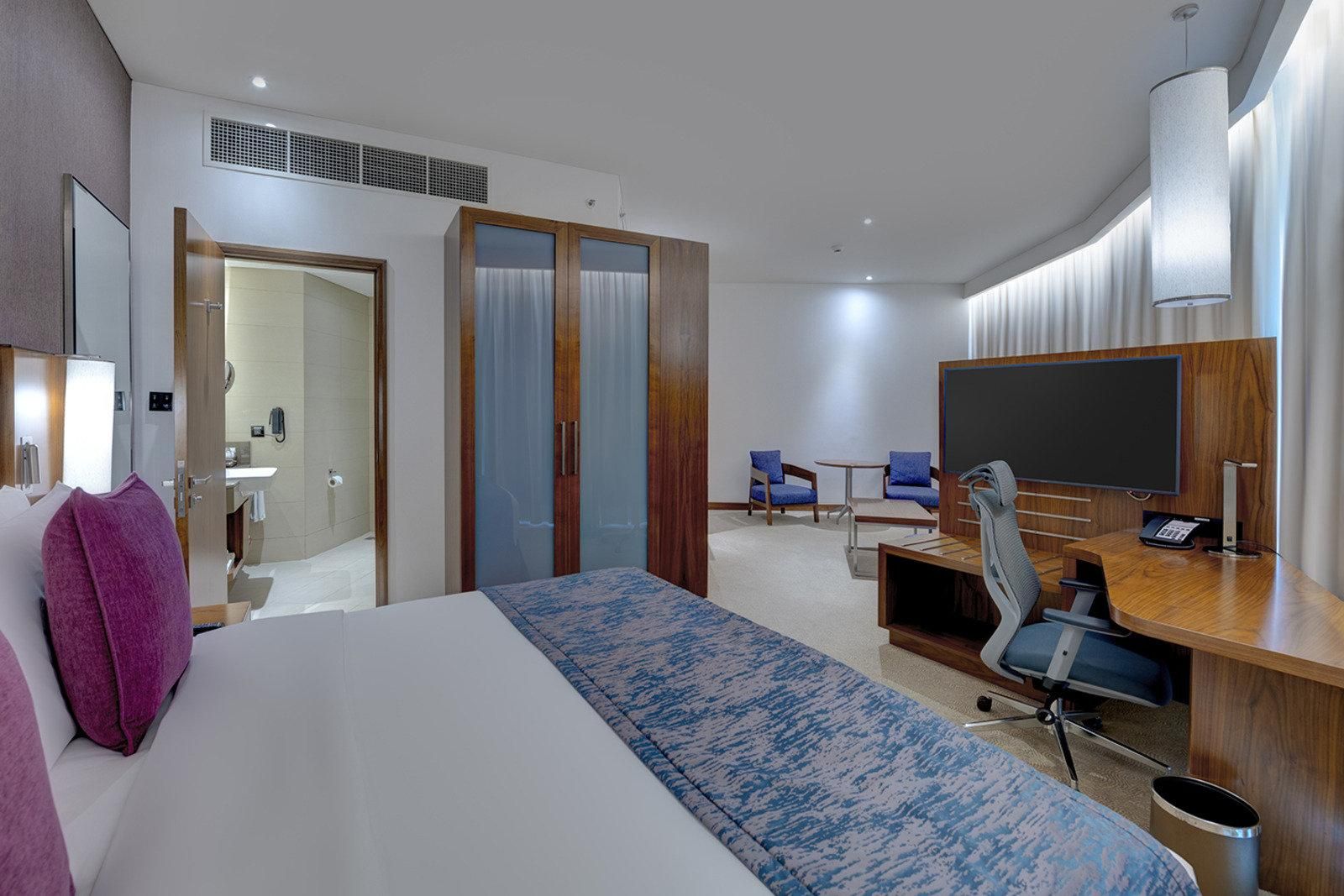 A hotel room with a bed, a desk with a chair, a television, and a bathroom visible through an open door.