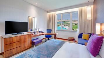 A hotel room with a large bed, a desk with a flat-screen TV, a chair, a small table, and a view of a pool and landscape.
