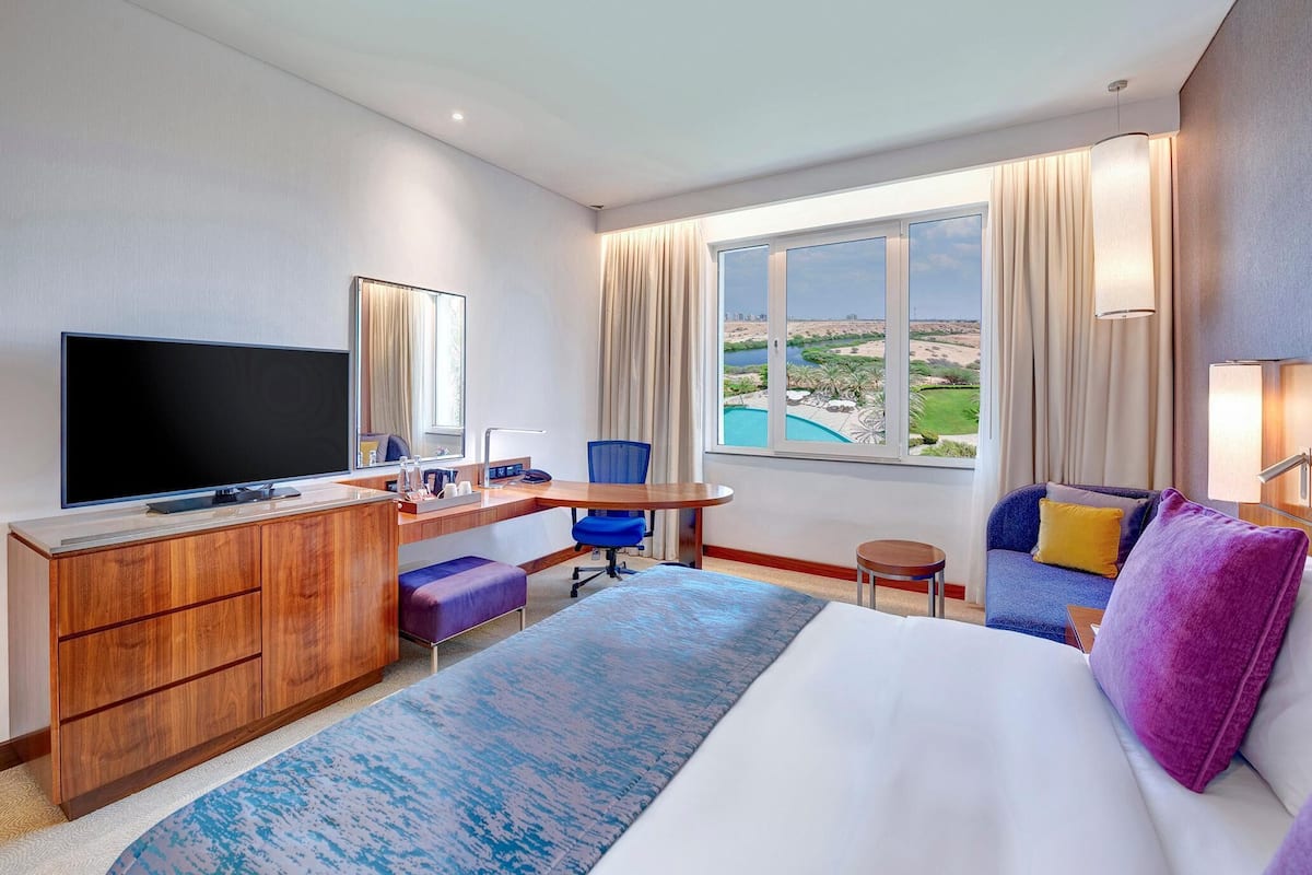Habitación de hotel con una cama grande, un escritorio con televisor, una silla, una mesita y vistas a una piscina y al paisaje.