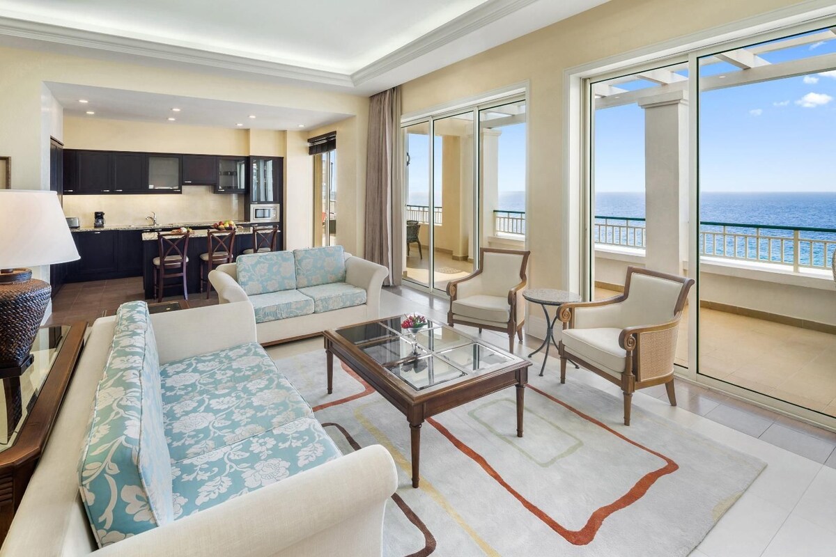 A spacious living room with a sofa, armchairs, and a coffee table. There is a kitchen area in the background and large windows with a view of the ocean.