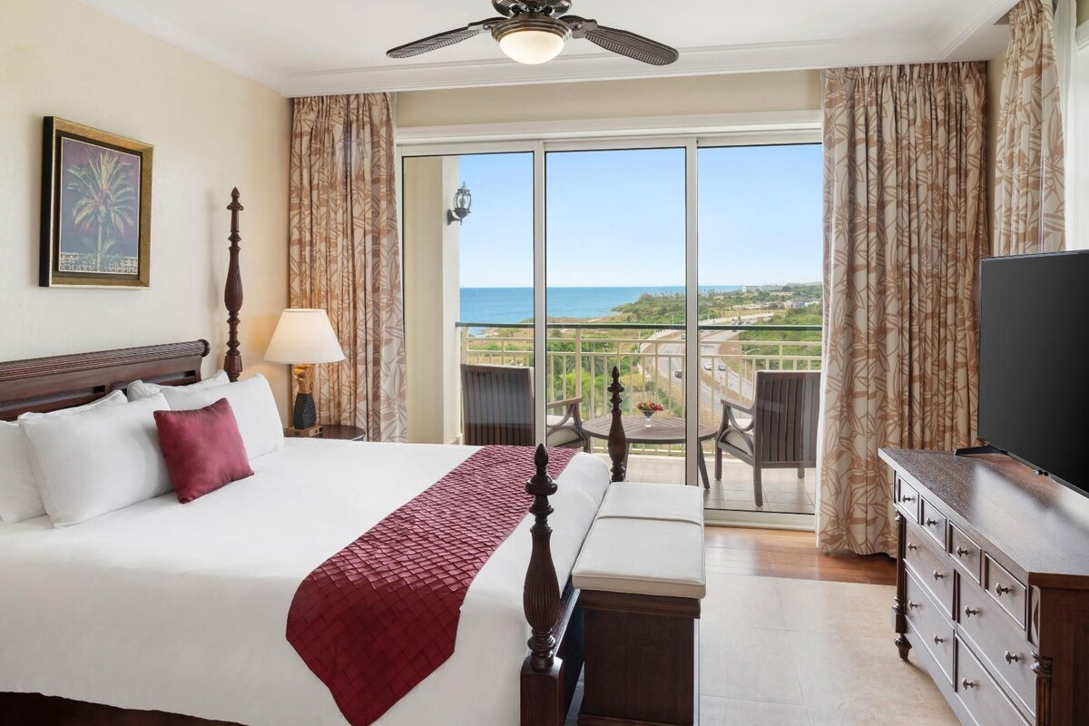 A hotel room with a bed, a TV, a balcony with a view of the ocean, and a ceiling fan.